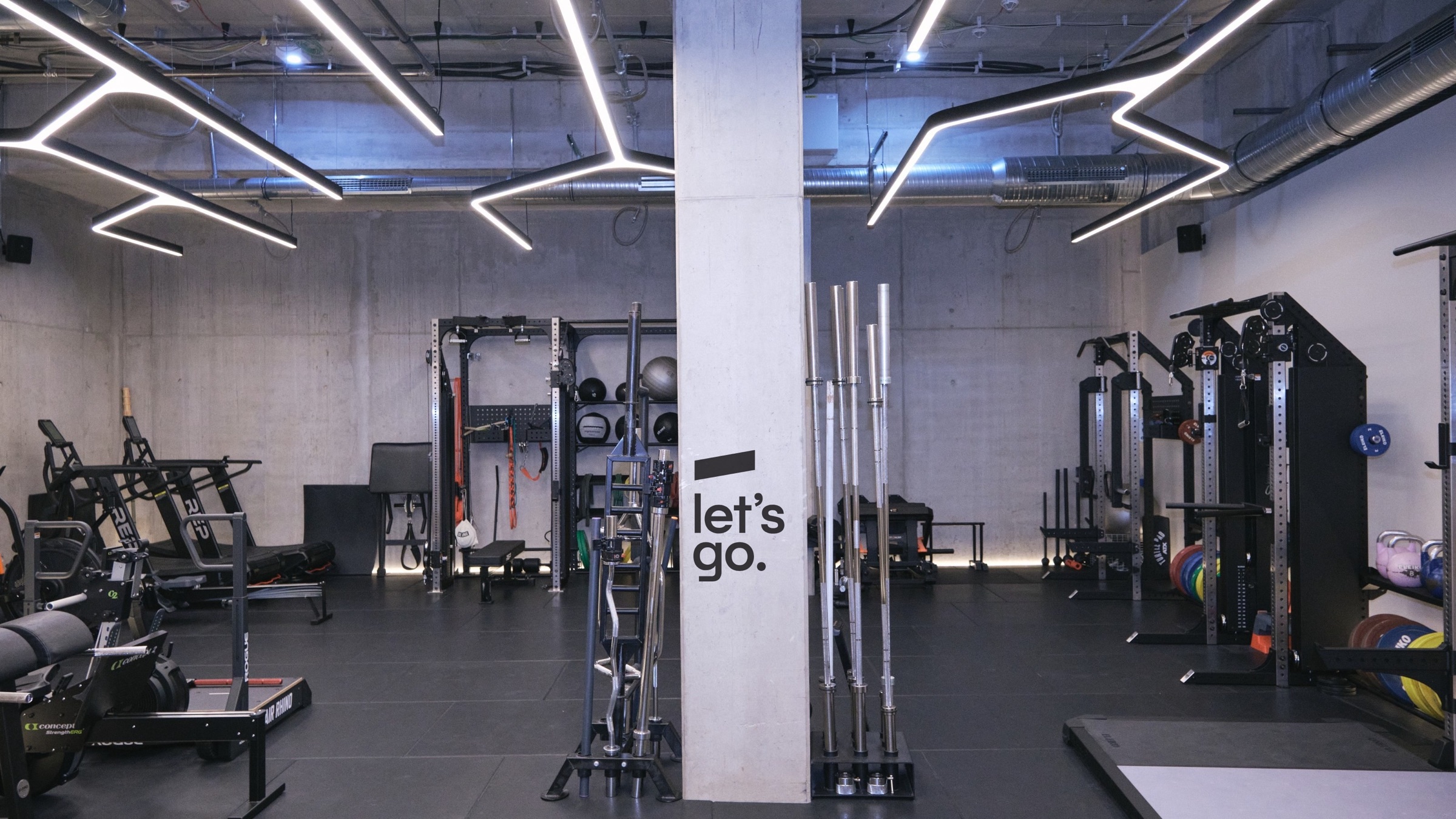 Trainingsfläche im neuen Studio mit Racks, Langhanteln und Geräten vor Sichtbetonwänden, auf der Säule der Schriftzug let’s go.